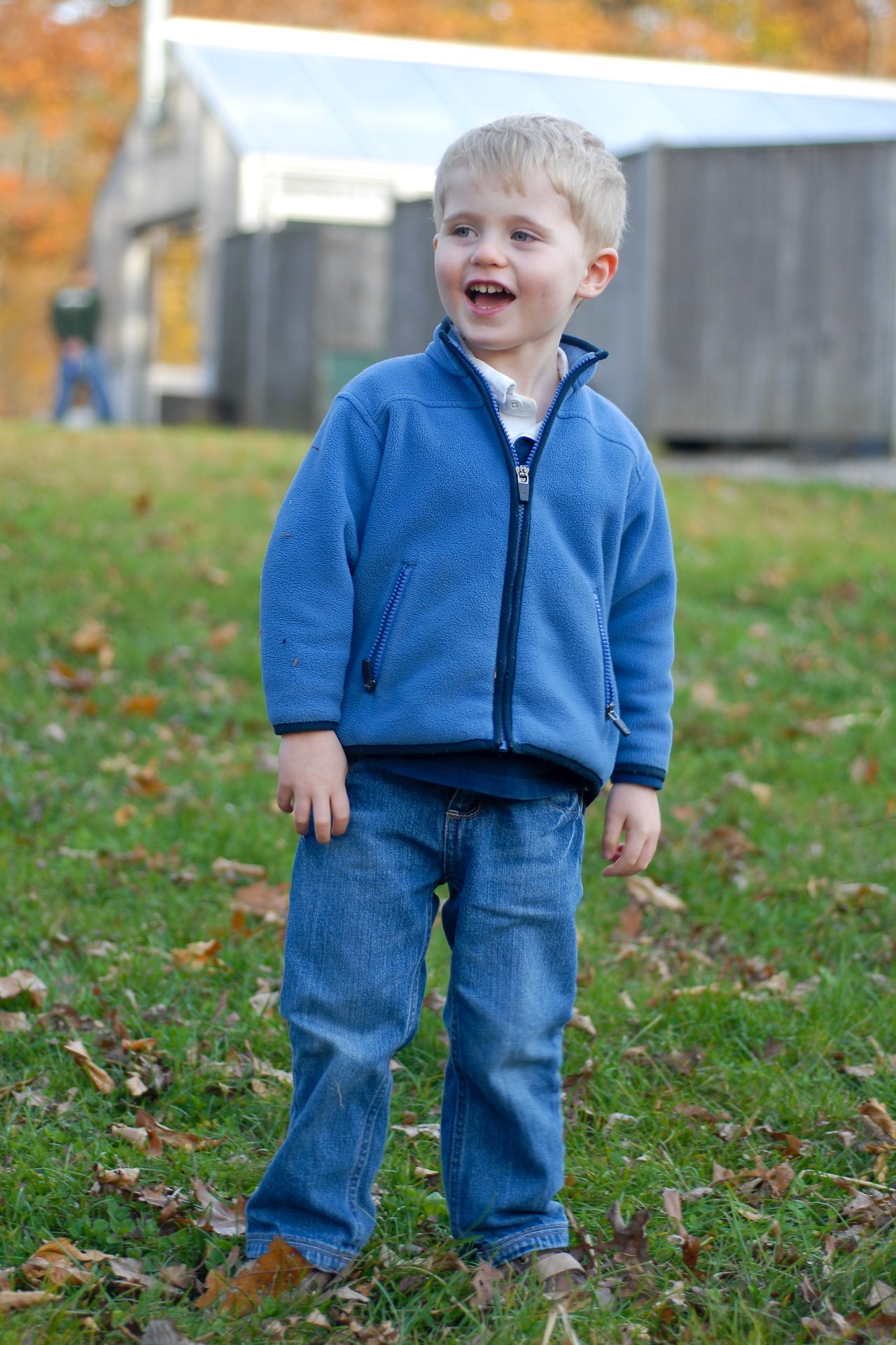 A young child in a blue jacket stands on grass, smiling and looking off to the side.