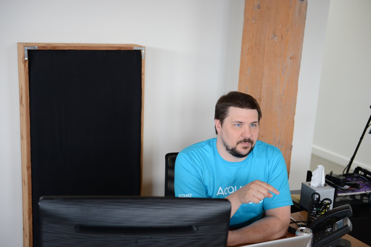 A man in an Acquia t-shirt sits at a desk, pointing while looking at a computer screen.