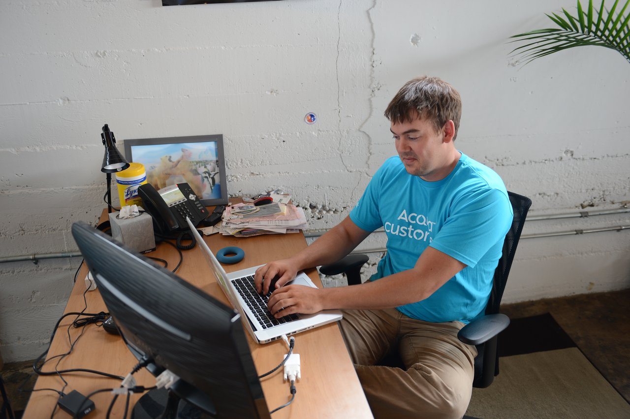 A man in an Acquia t-shirt types on a laptop at his desk in an office.