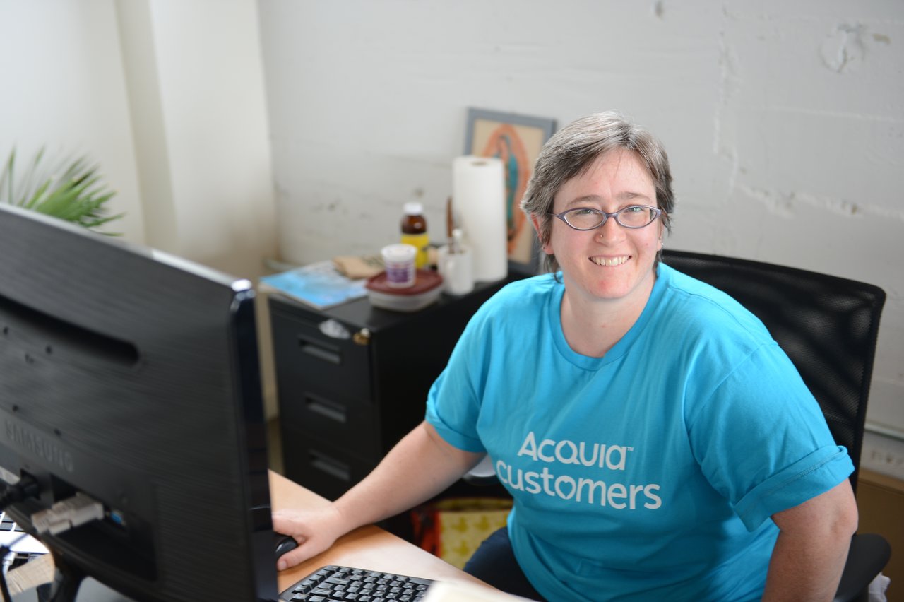 A person wearing an "Acquia Customers" shirt sits at a desk, smiling while working on a computer.
