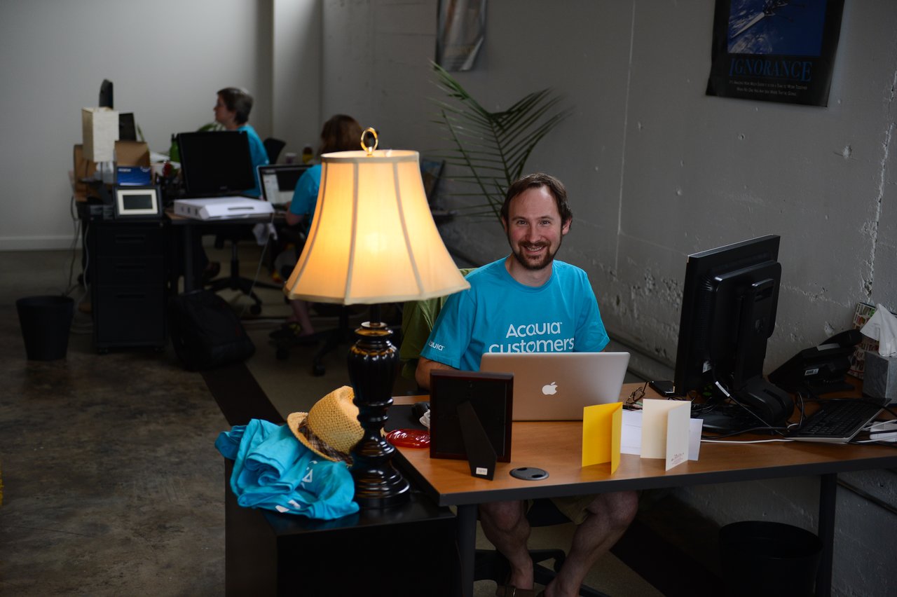 A man in an Acquia t-shirt smiles while working on a laptop at a desk in an office.