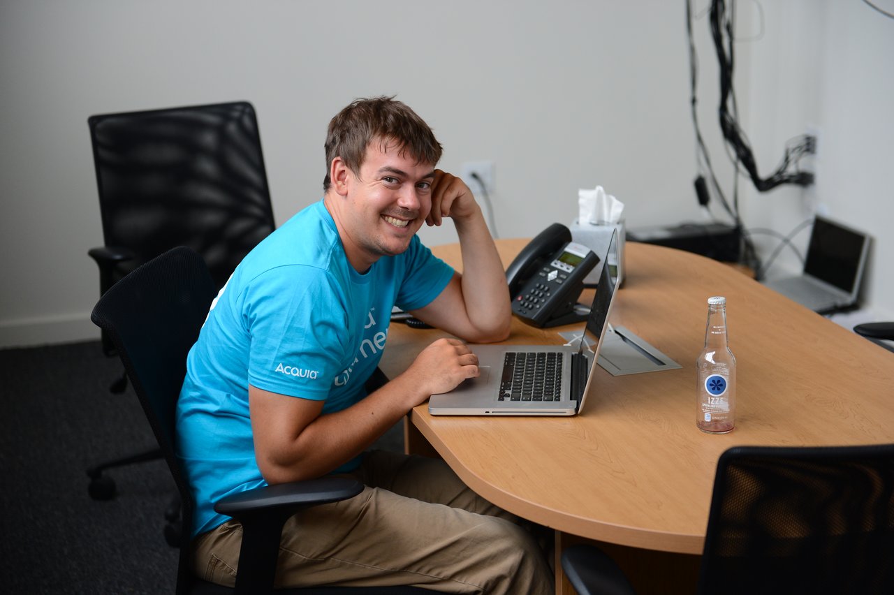 A person in a blue Acquia shirt sits at a desk, smiling while working on a laptop.