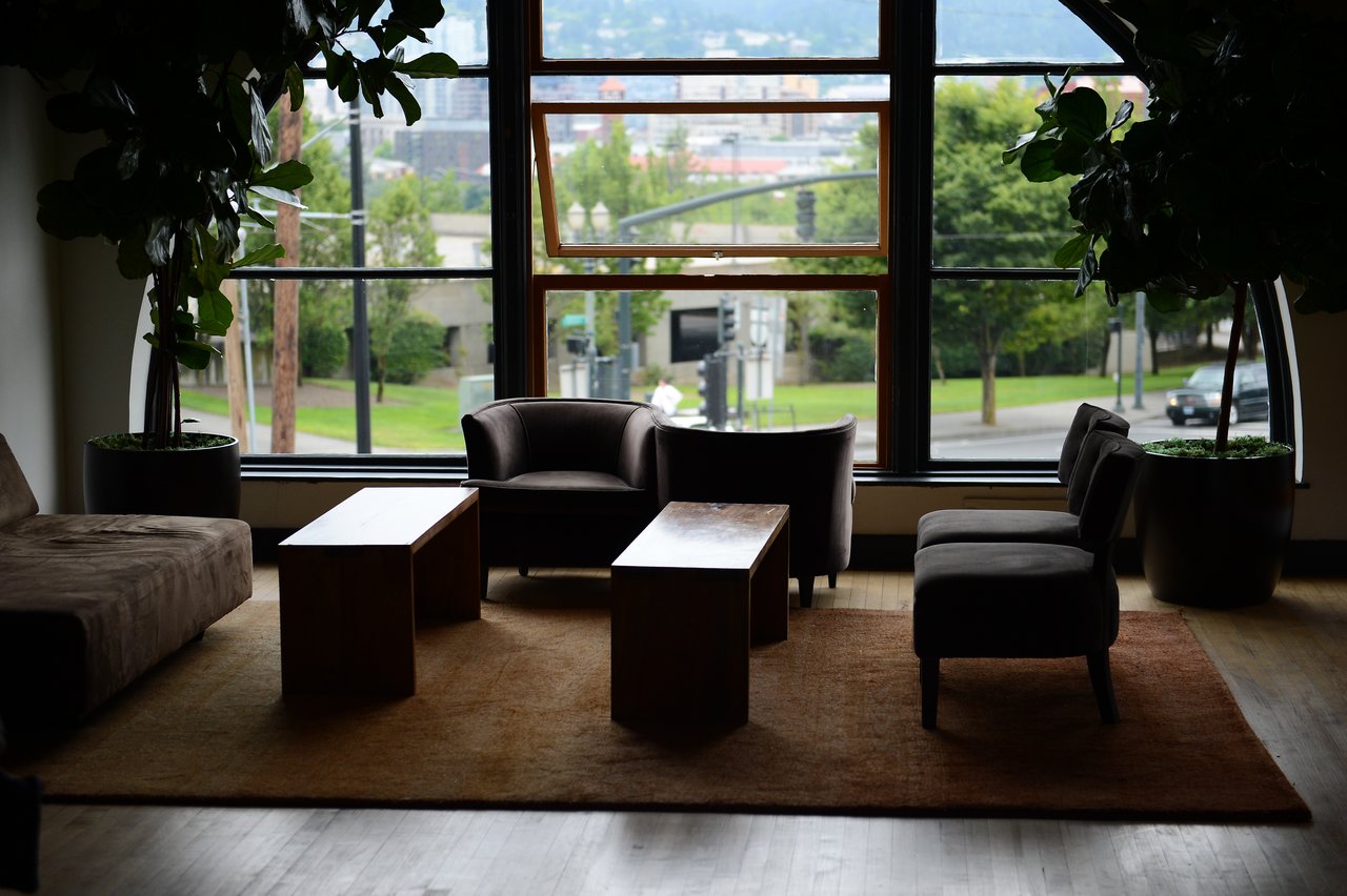 A modern office lounge with chairs, tables, and large windows overlooking a city street with greenery.
