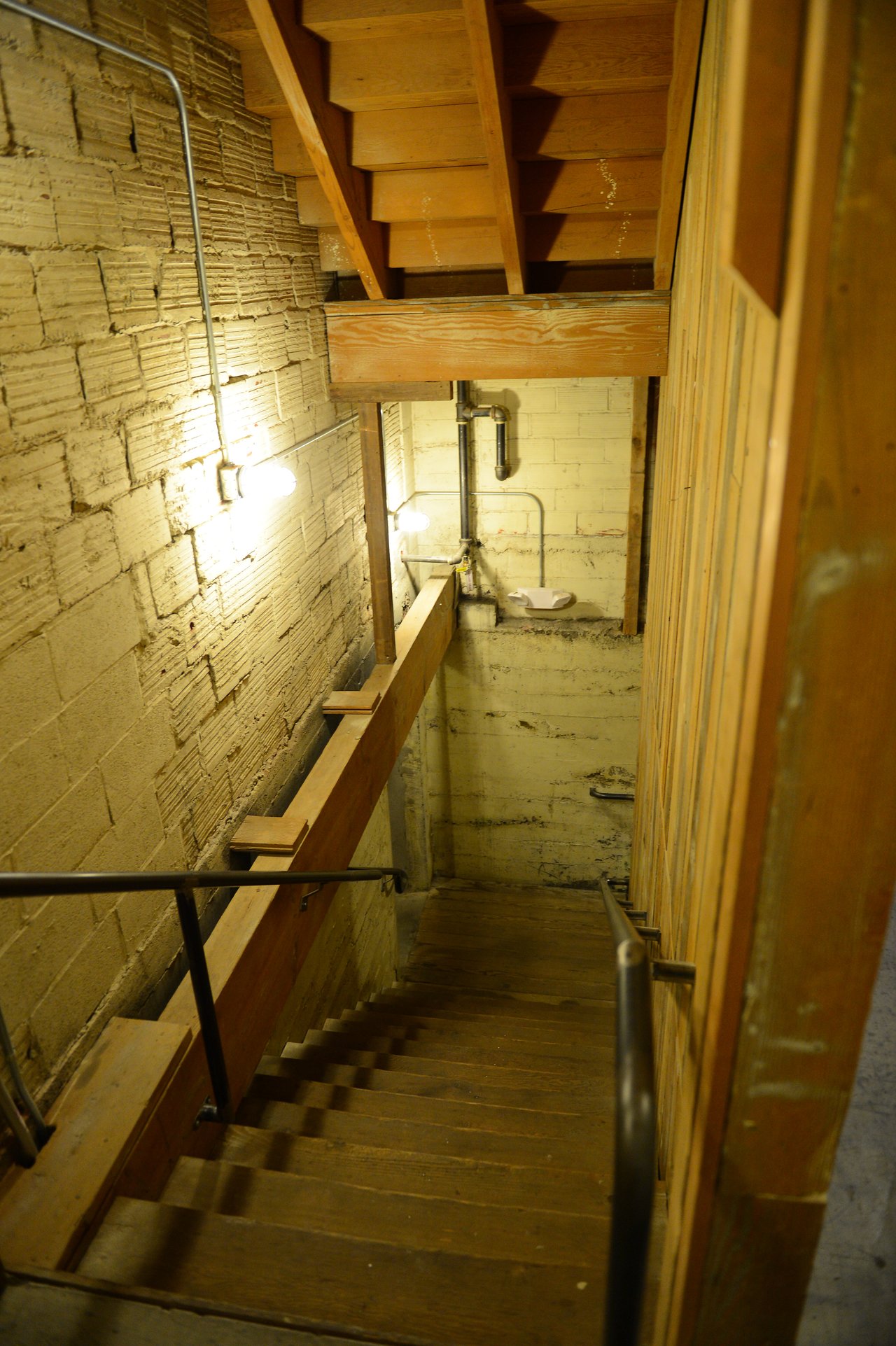 A wooden staircase with metal railings leads down to a basement area with exposed brick and concrete walls.