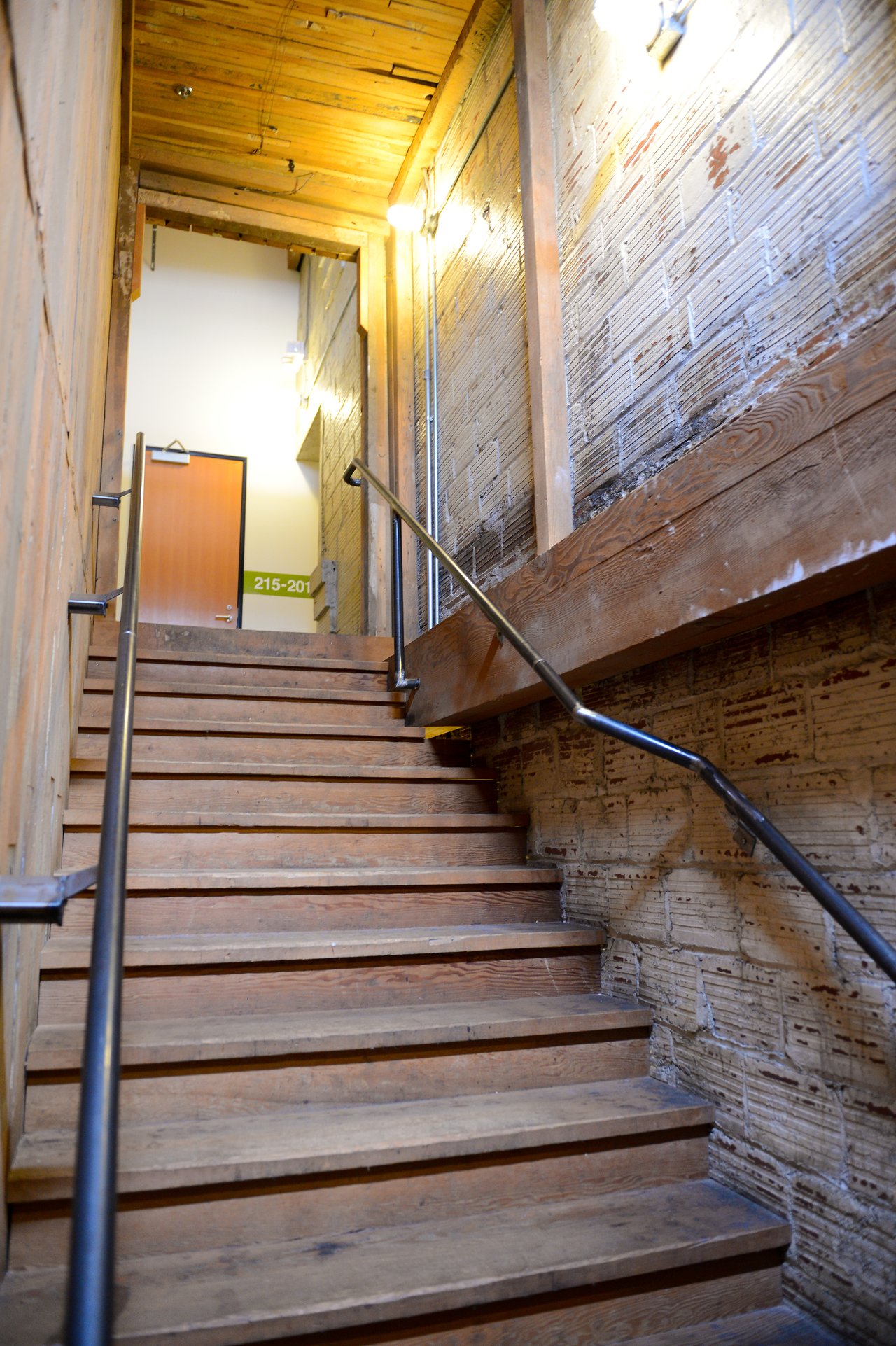 A wooden staircase with metal handrails leads to an office door with a room number sign.