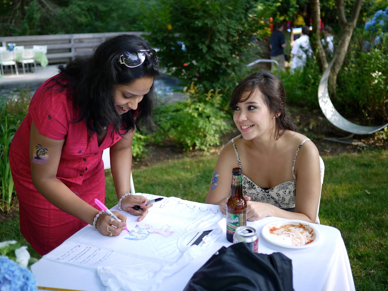 A woman in a red dress signs a white T-shirt while another woman smiles and watches at an outdoor party.