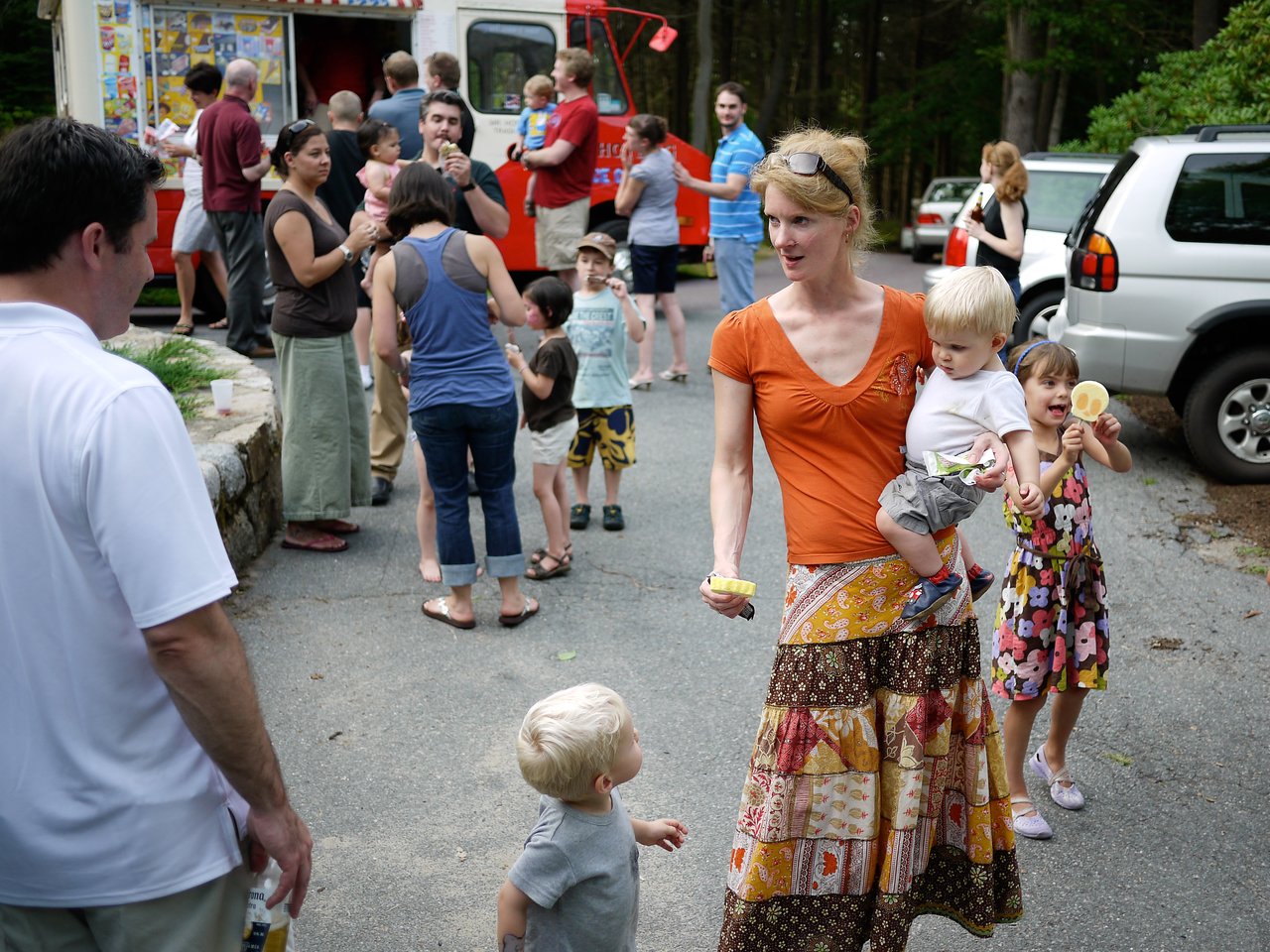 A woman carrying a child walks with two children holding ice cream, while people gather near an ice cream truck.