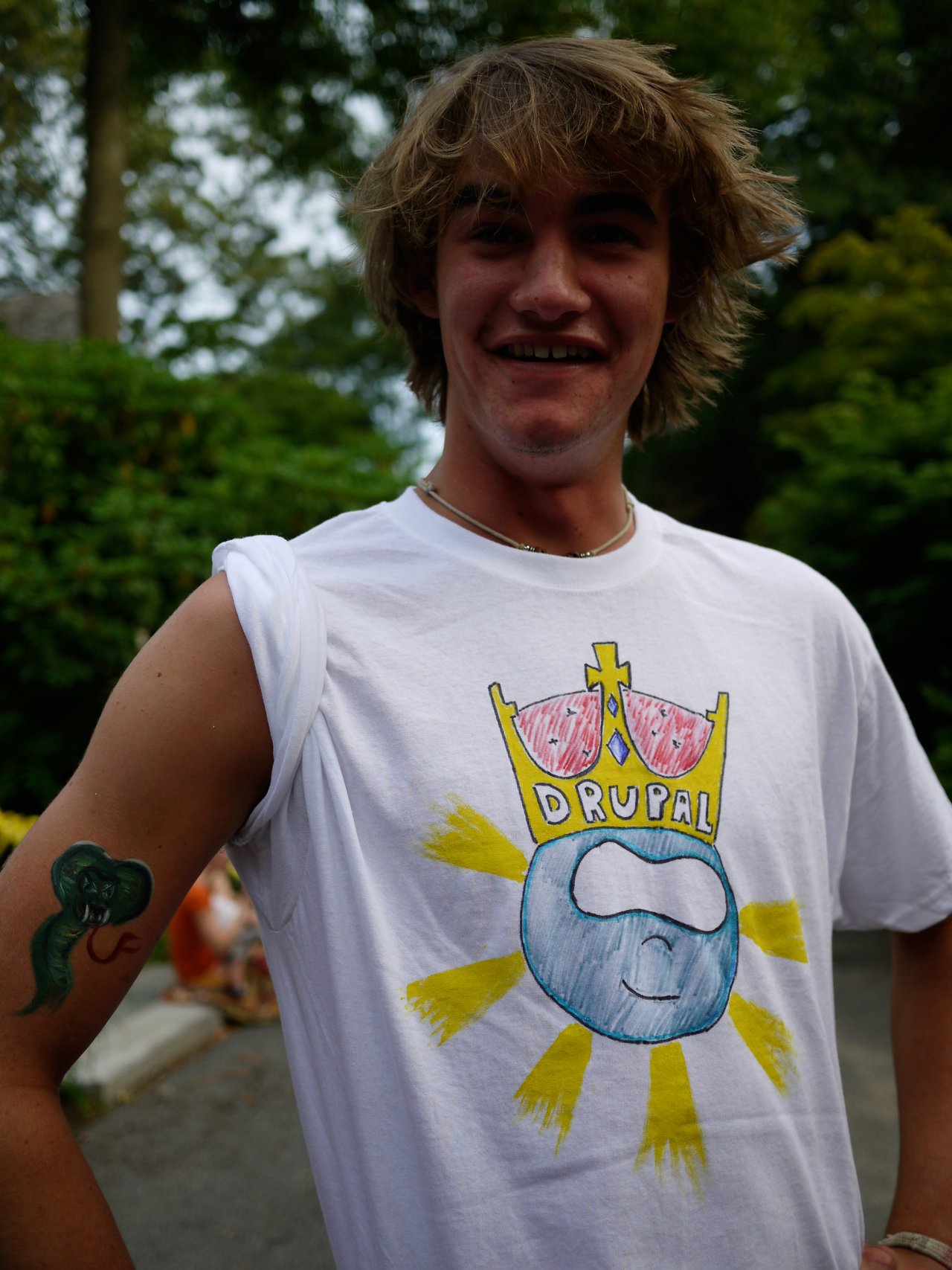 A smiling person wears a white T-shirt with a hand-drawn Drupal logo and has a temporary tattoo on their arm.