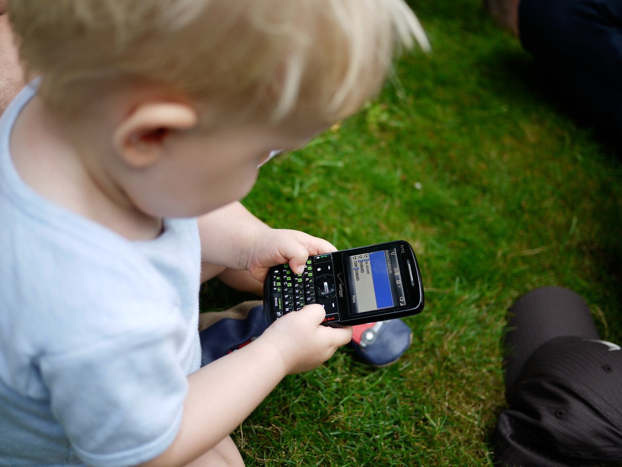 A young child sits on the grass, holding a smartphone and looking at the screen.