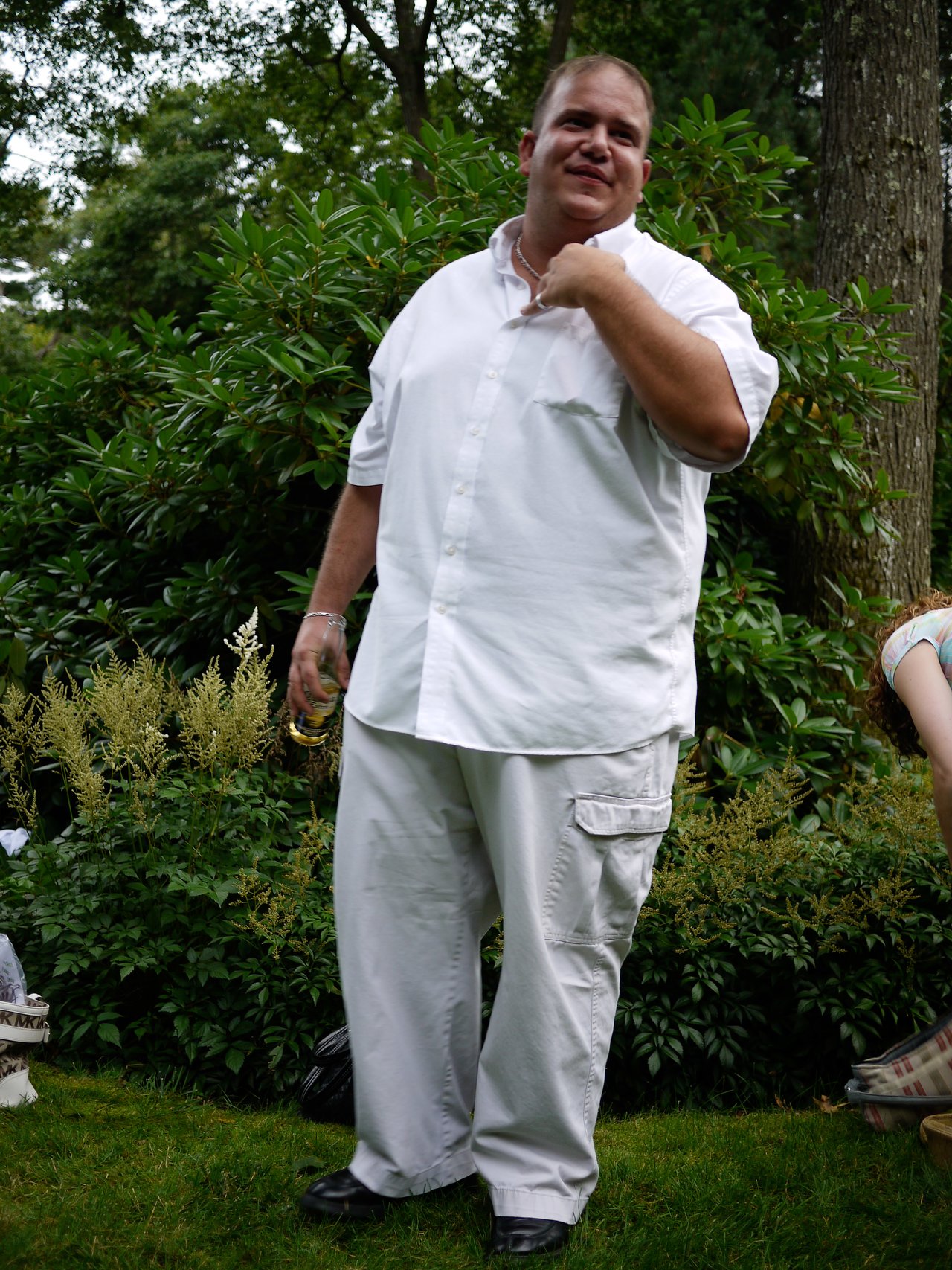 A man in a white outfit stands outdoors, holding a drink and touching his neck while smiling.