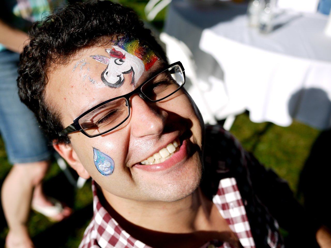 A smiling person with glasses and face paint, including a unicorn on their forehead, at an outdoor party.