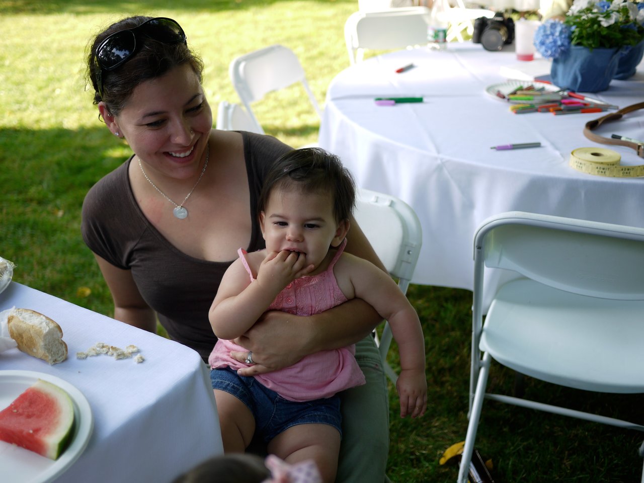 A smiling woman holds a young child who is eating with her fingers at an outdoor party with tables and chairs.