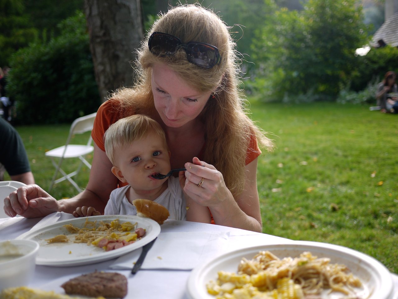 A woman feeds a young child at an outdoor gathering, with plates of food on the table.