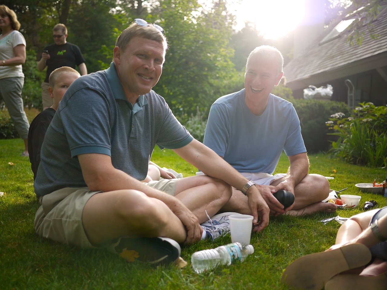 Two men sit on the grass, smiling and chatting at an outdoor gathering with food and drinks nearby.