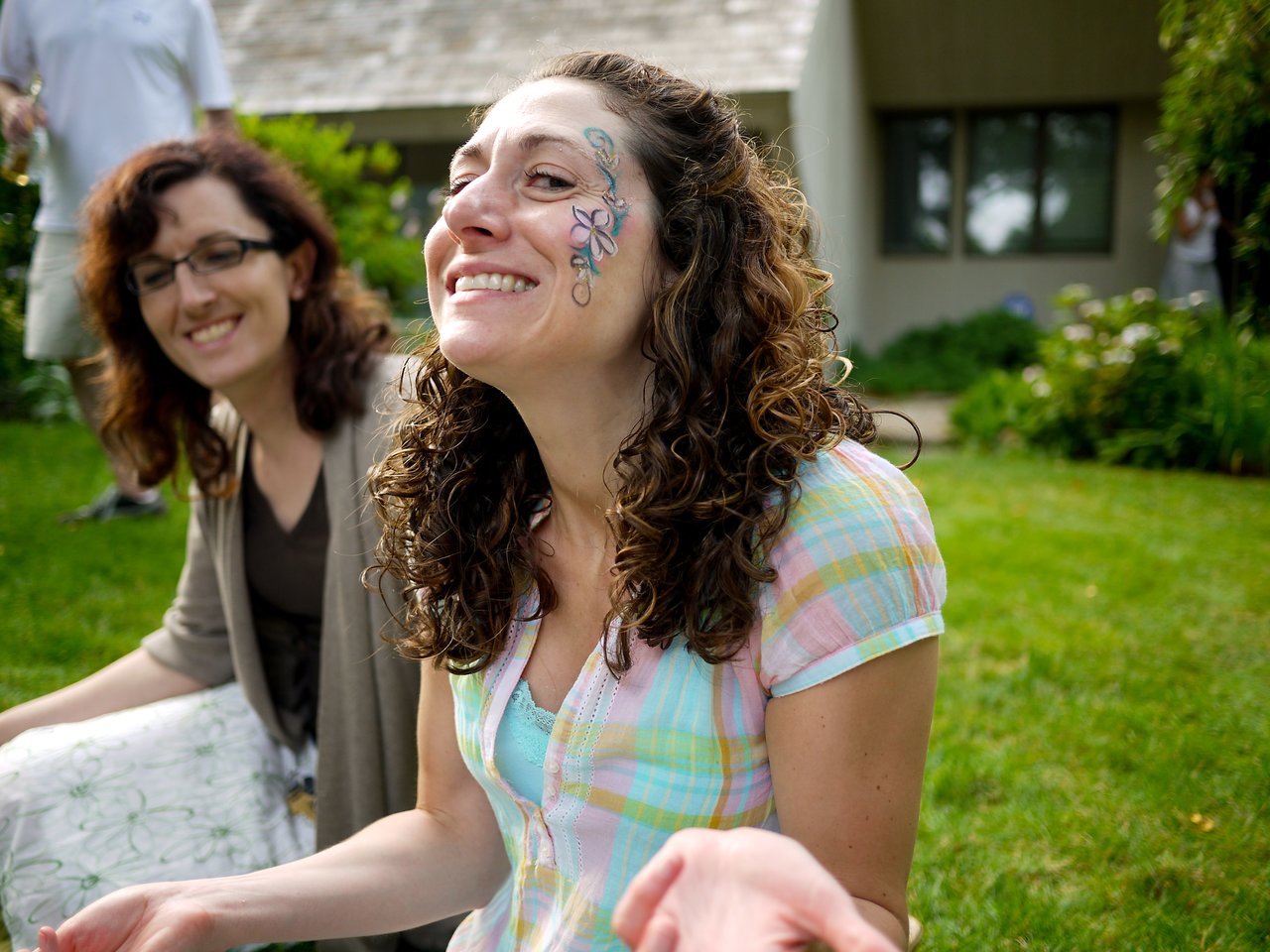 Two smiling women sit on the grass, one with face paint, enjoying an outdoor gathering.