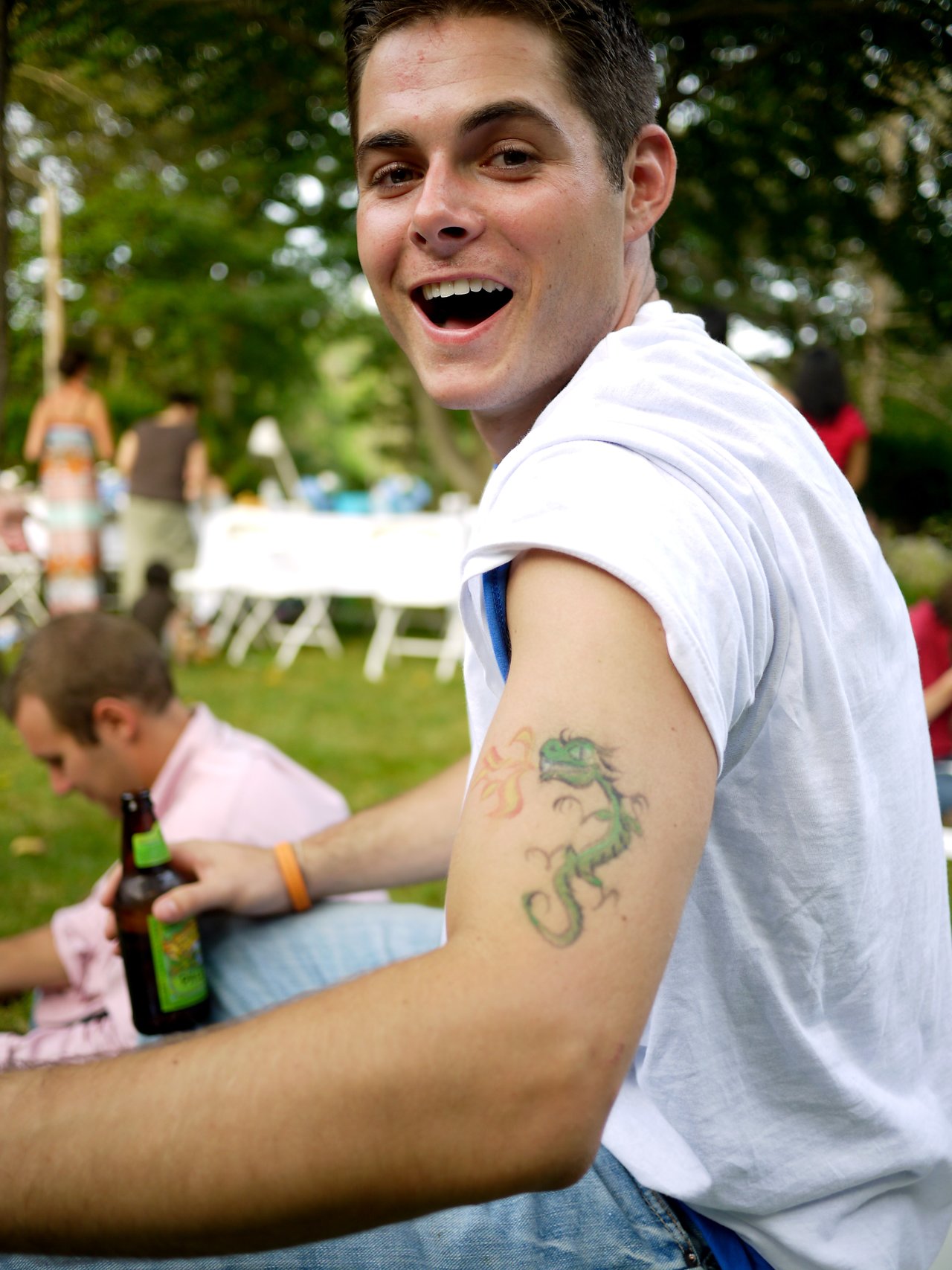 A smiling man in a white shirt shows off a dragon tattoo on his arm at an outdoor party.
