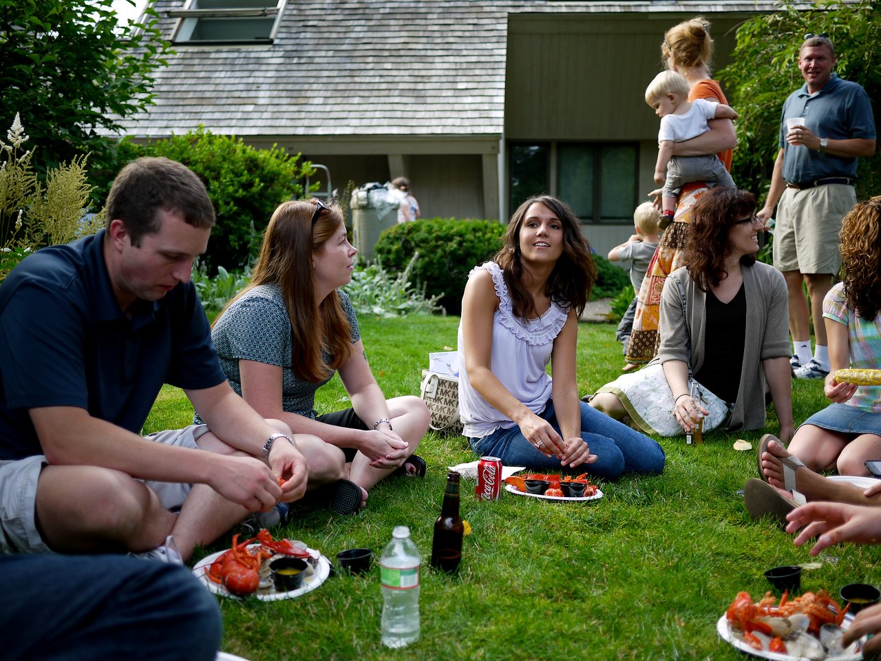 People sit on the grass, eating lobster and talking at an outdoor summer party.