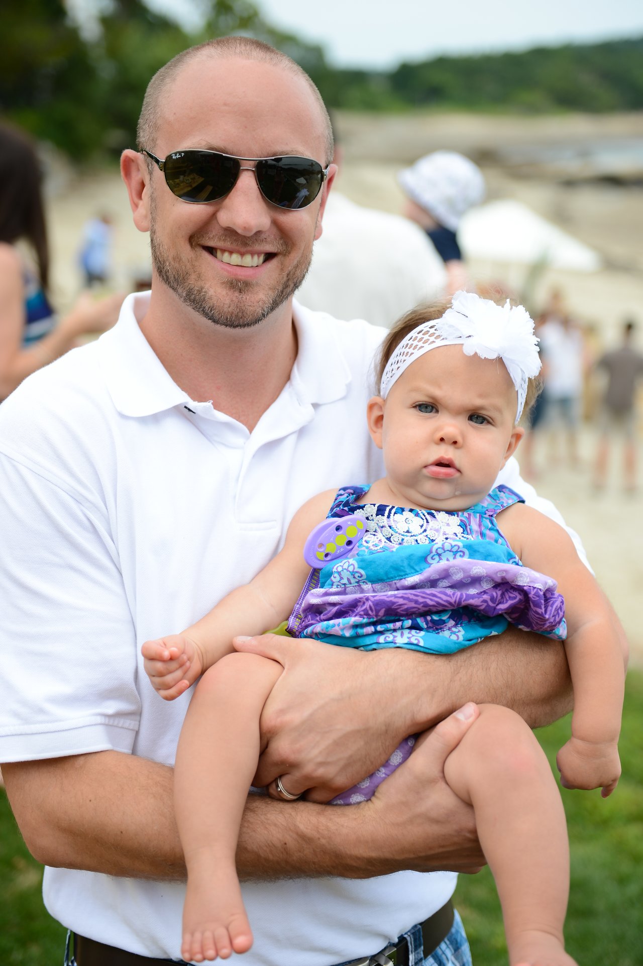A man wearing sunglasses and a white shirt smiles while holding a baby dressed in blue and purple.