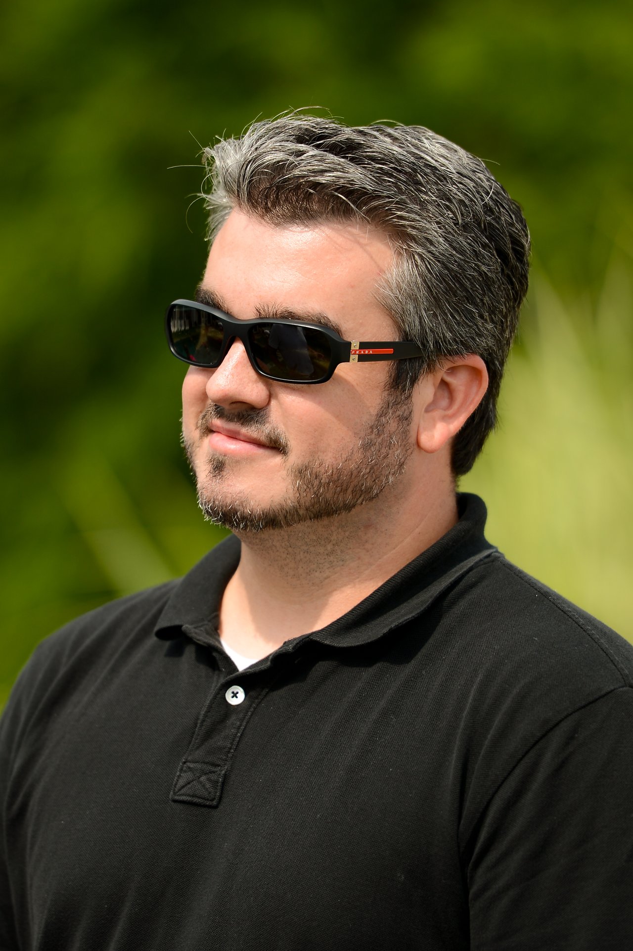 A man wearing sunglasses and a black polo shirt stands outdoors, looking off to the side.