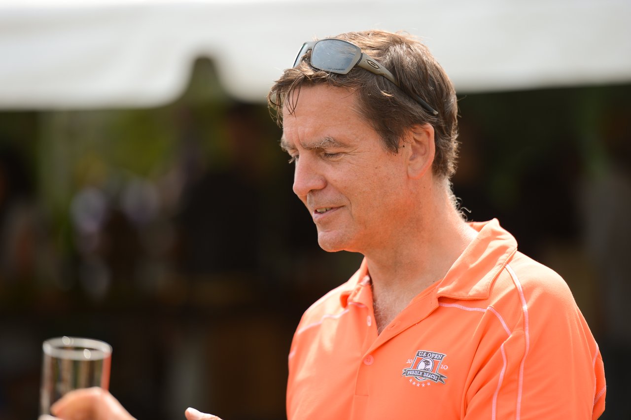 A man in an orange polo shirt holds a drink and talks at an outdoor party.