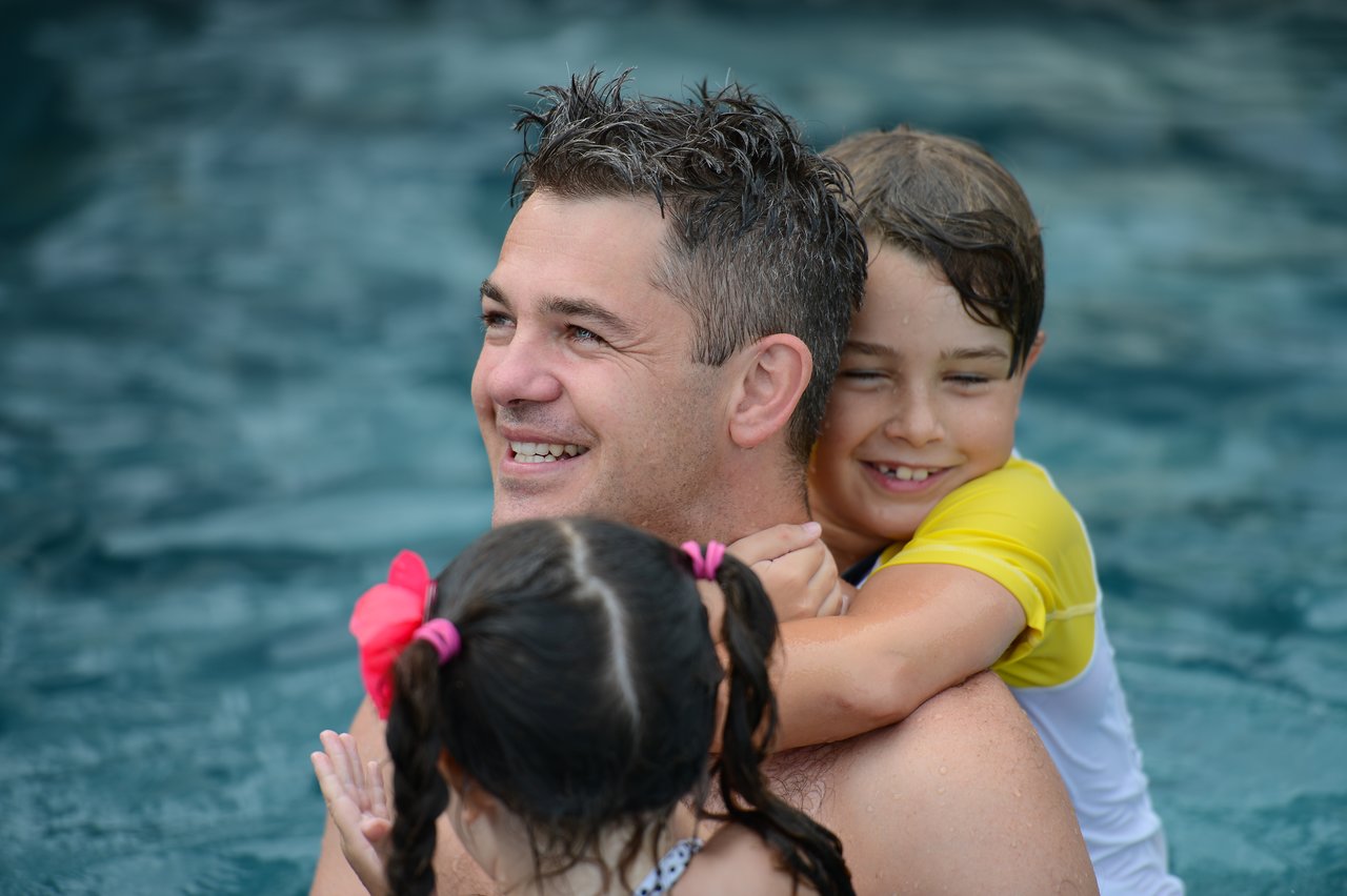 A man in a pool smiles while two children hug him from behind, enjoying the water together.