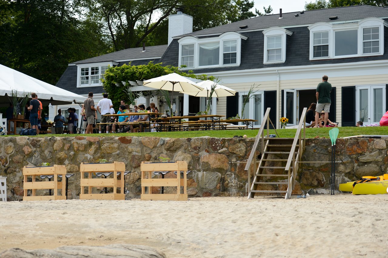 People gather at picnic tables and under a tent outside a large house for a summer party.