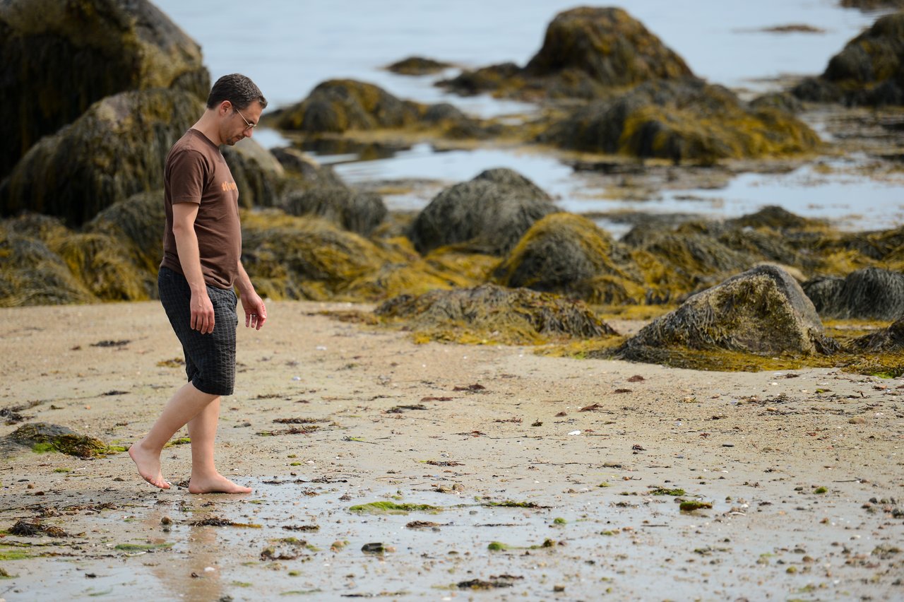 A person walks barefoot on a rocky beach, looking down at the sand and water.