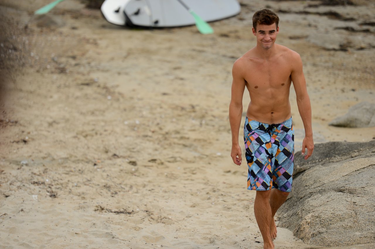 A man in colorful swim trunks walks barefoot on a sandy beach, smiling.