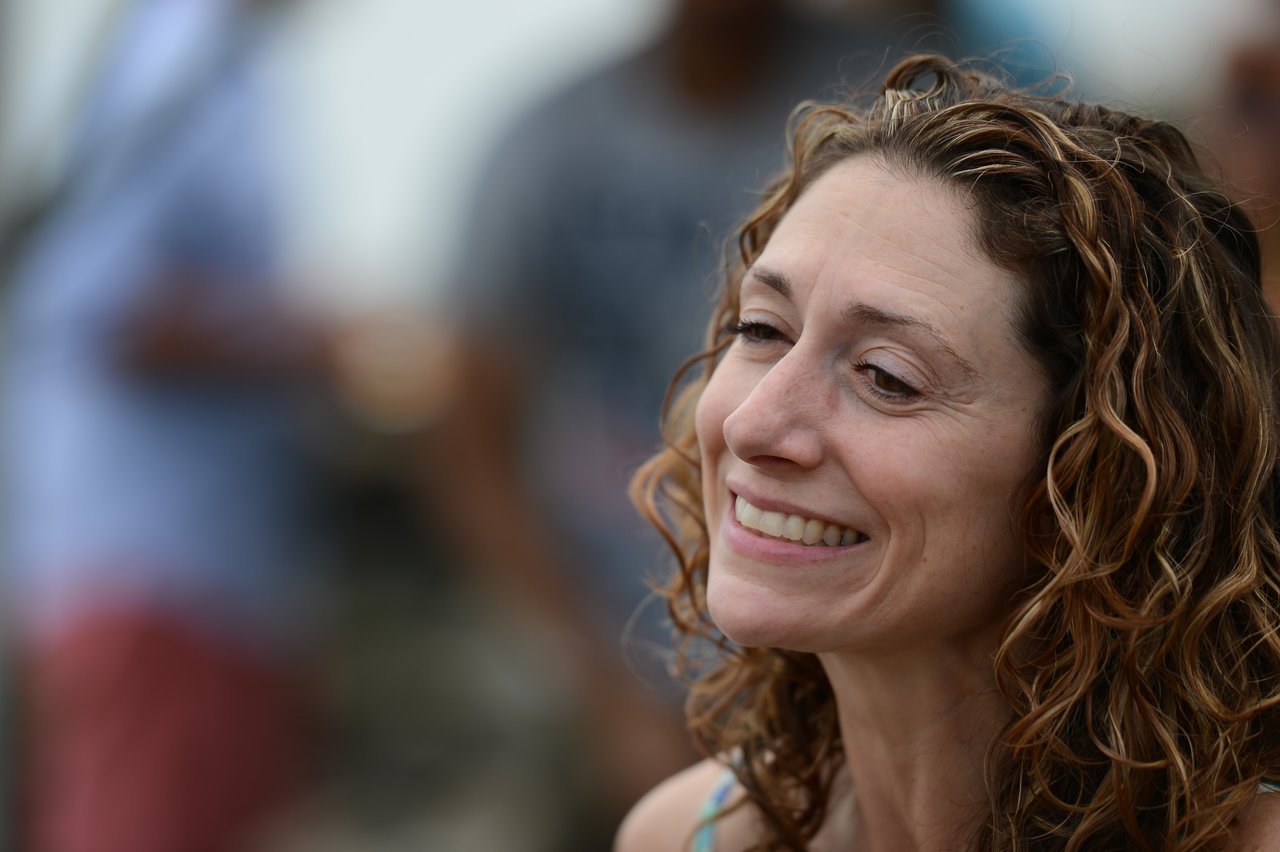 A woman with curly hair smiles while looking off to the side at an outdoor gathering.