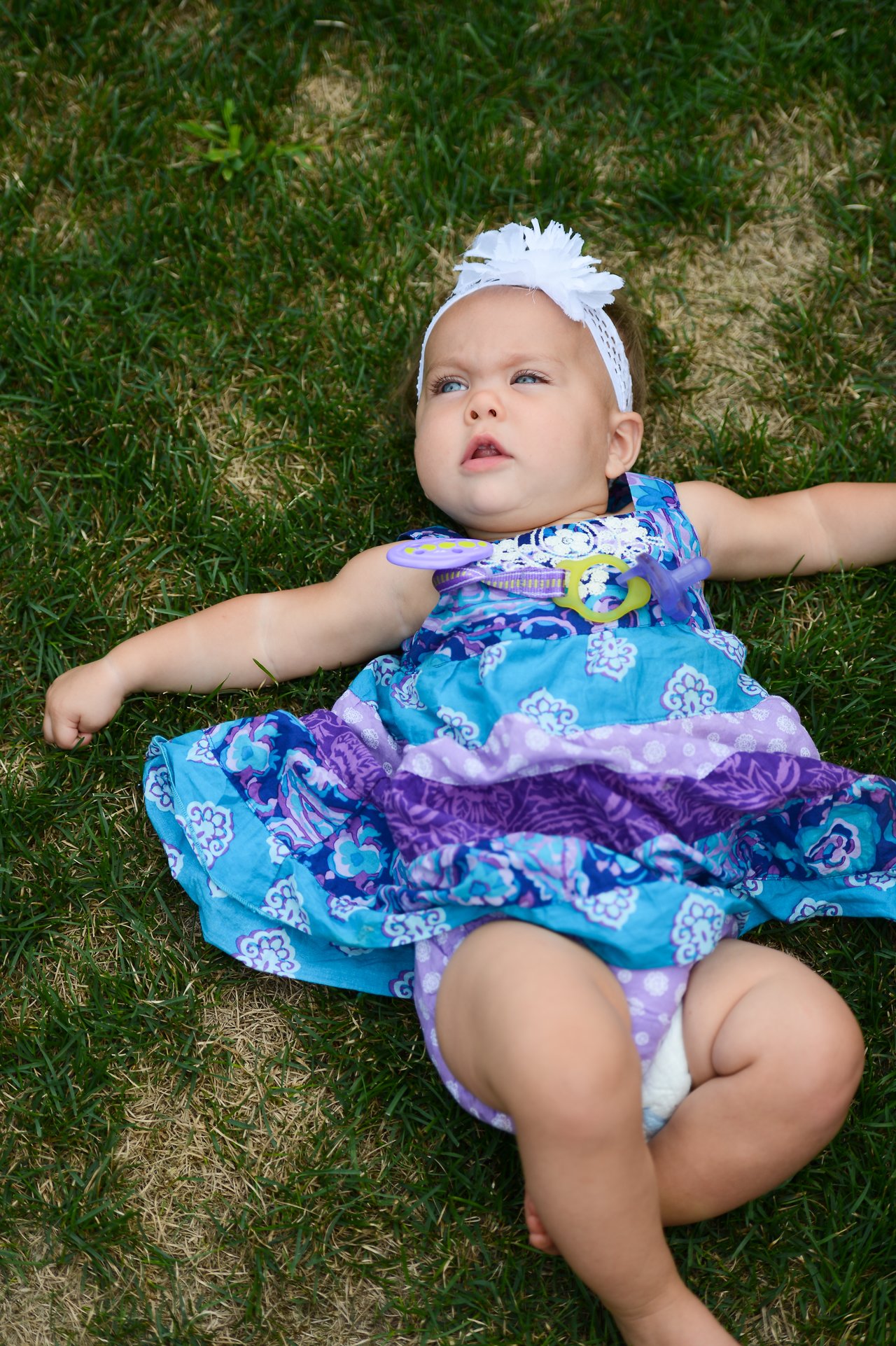 A baby in a colorful dress lies on the grass, looking up with arms outstretched.