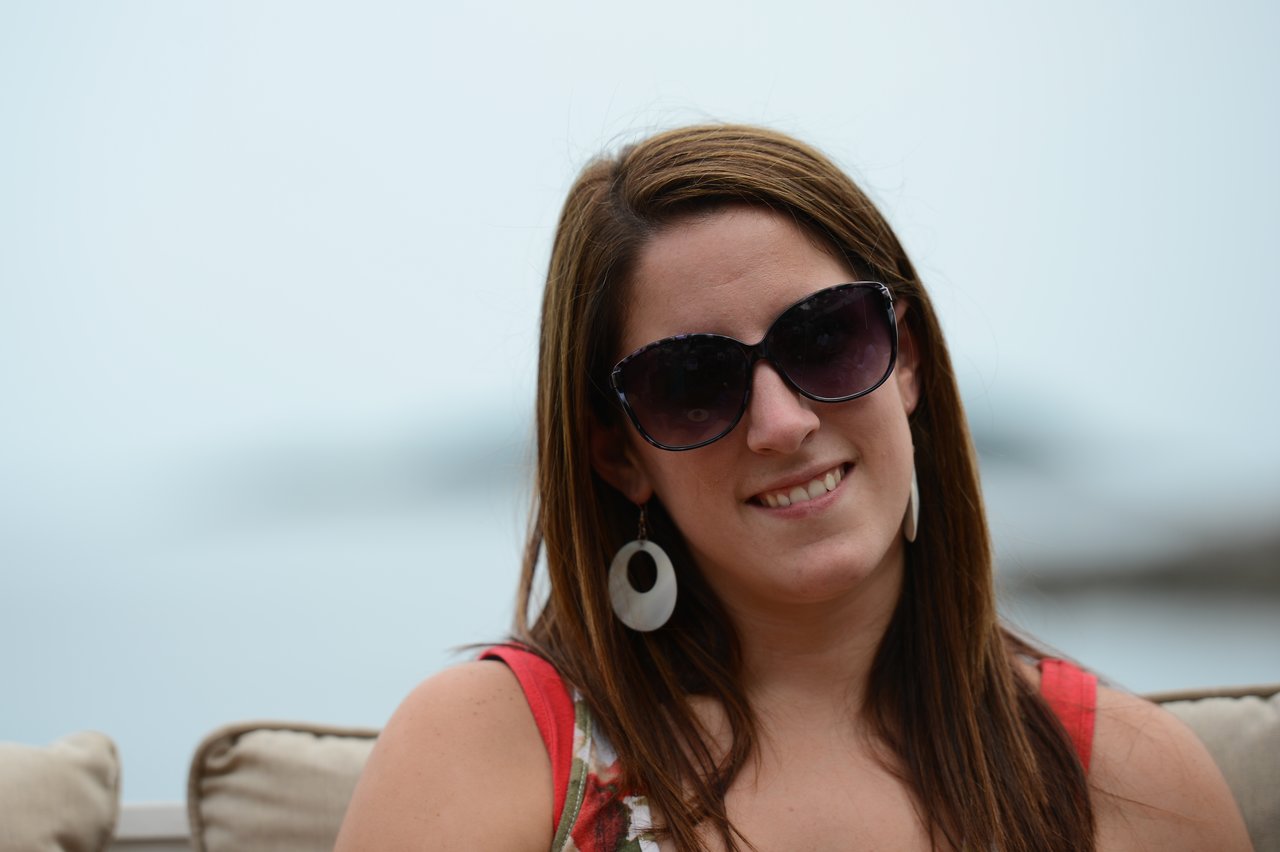 A woman wearing sunglasses and earrings smiles while sitting on an outdoor couch.