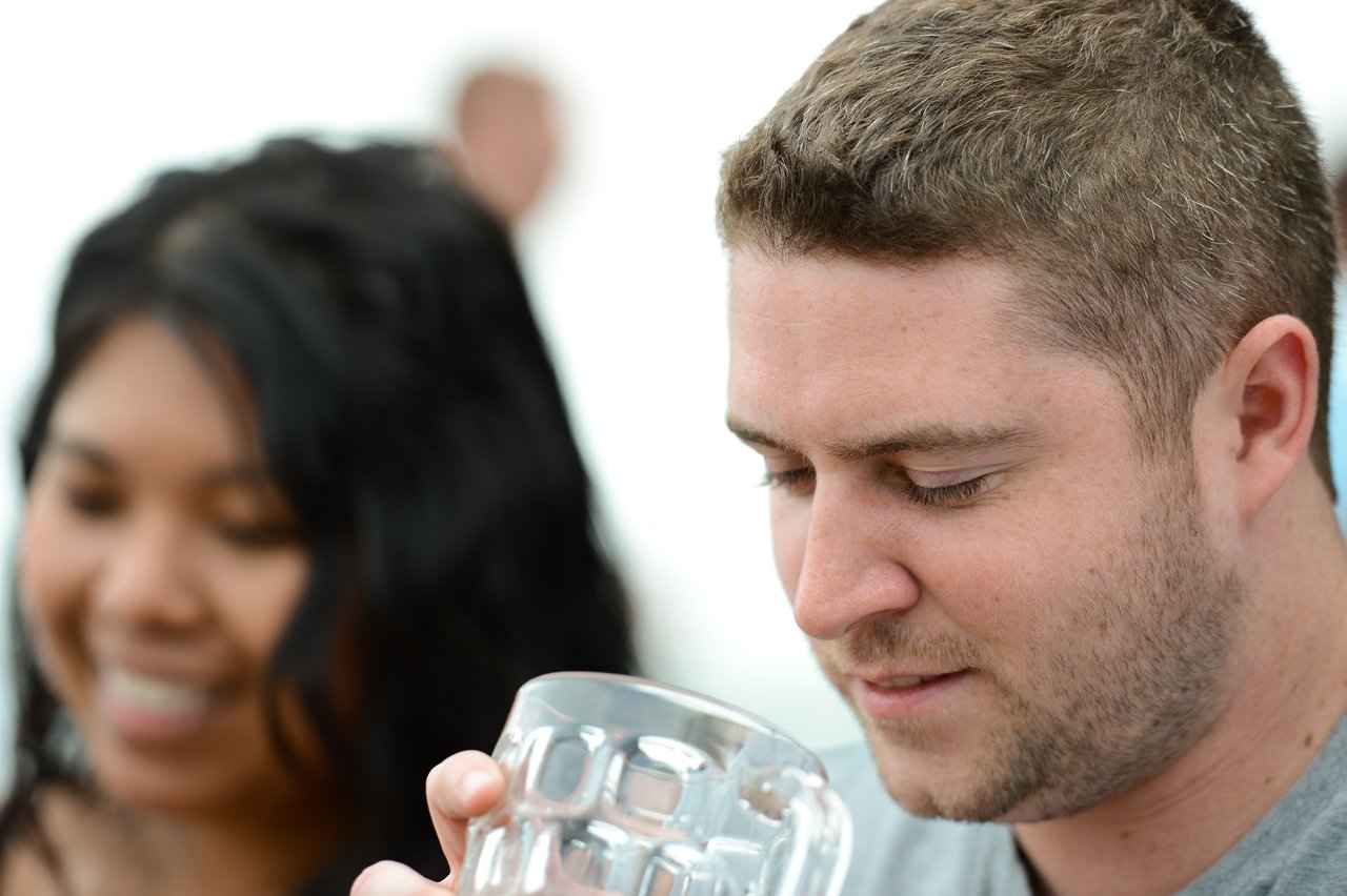 A man looks at his glass while a woman in the background smiles.