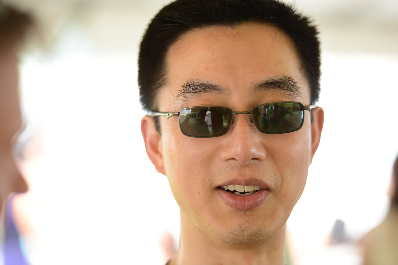A man wearing sunglasses is talking at an outdoor party, with reflections of people visible in his lenses.