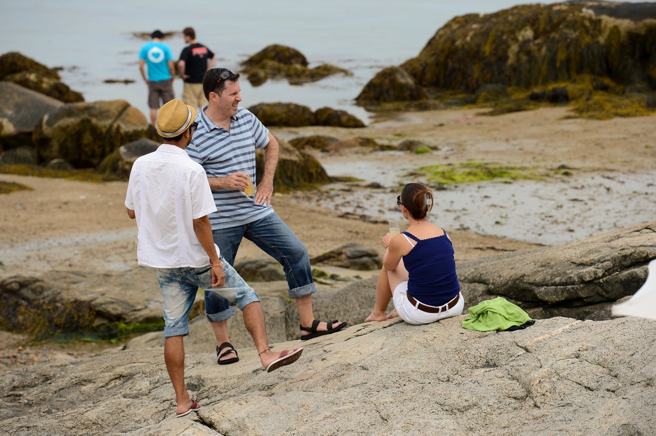 Three people are talking on a rocky beach, while two others stand near the water in the background.