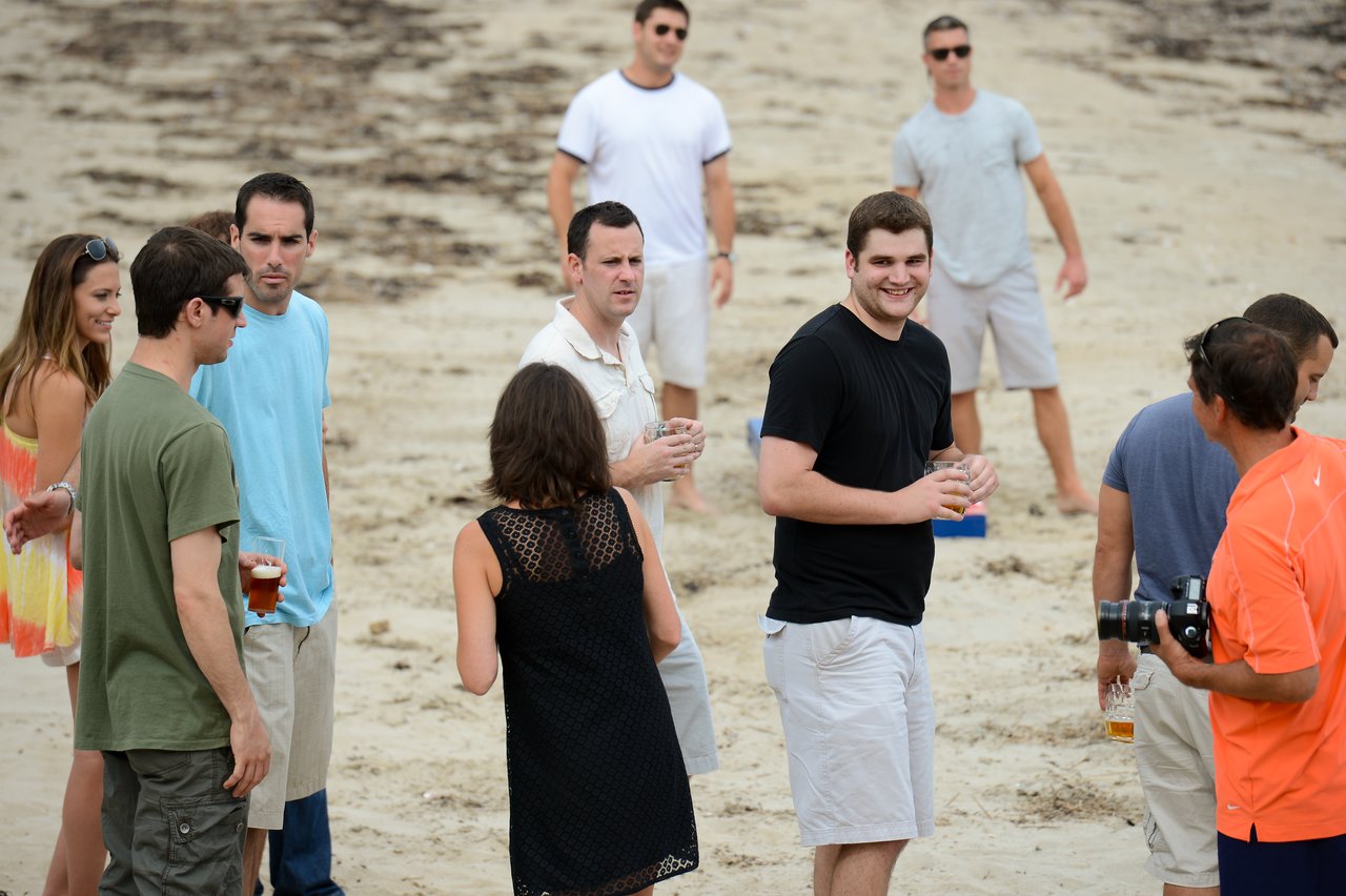 A group of people socializing on a sandy beach, some holding drinks and one person holding a camera.