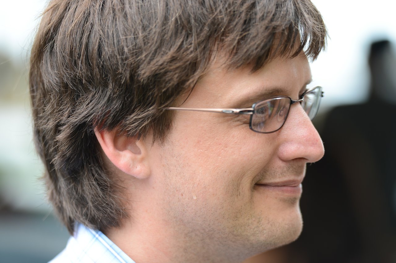 A man wearing glasses smiles while looking to the side at an outdoor gathering.