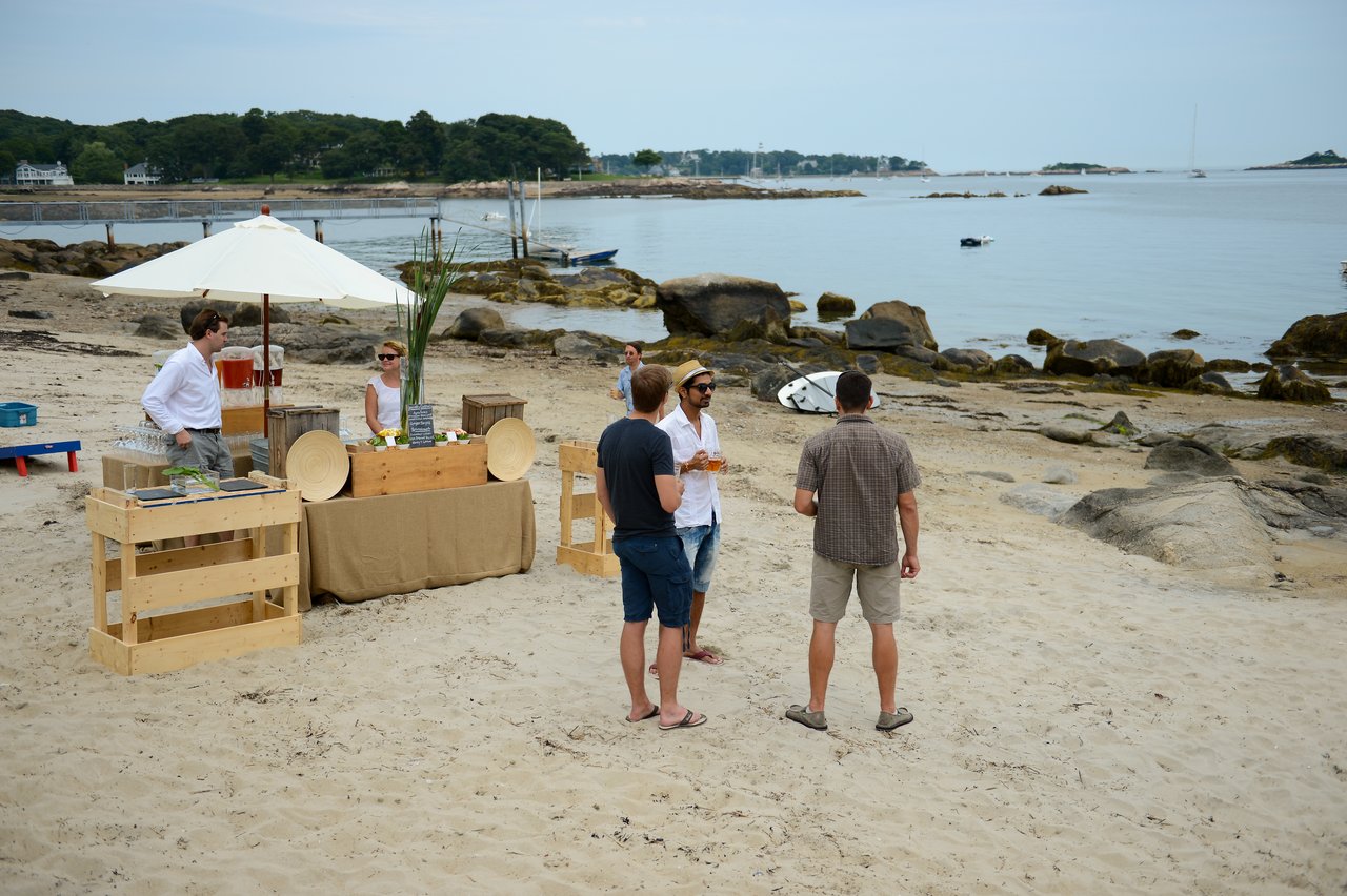 A small group of people socializing on a sandy beach near a refreshment stand with drinks and snacks.