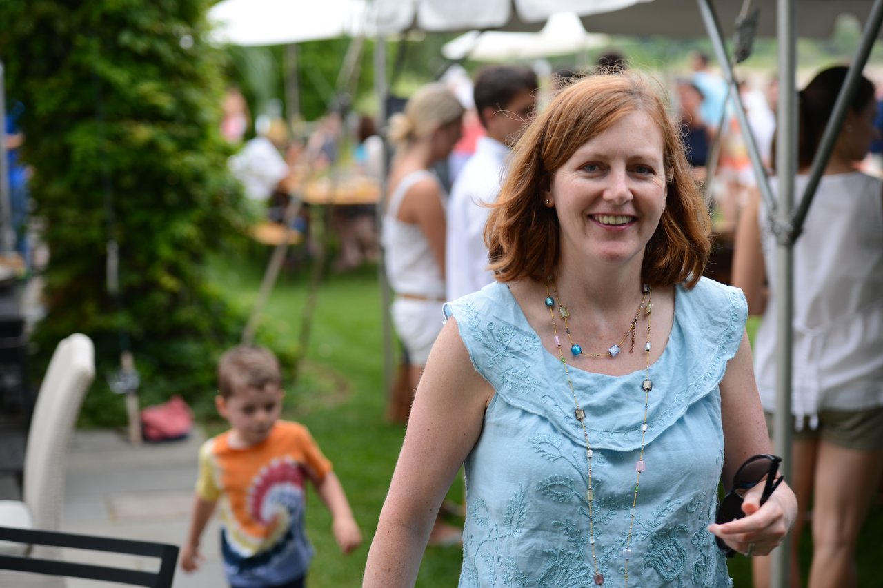 A smiling woman in a blue top walks toward the camera at an outdoor party, holding sunglasses in her hand.