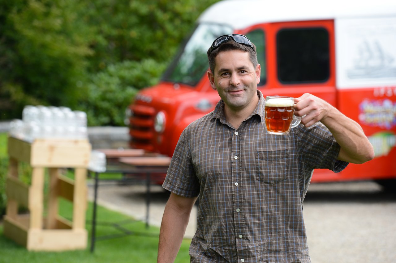 A man smiles while holding up a glass of beer at an outdoor party.