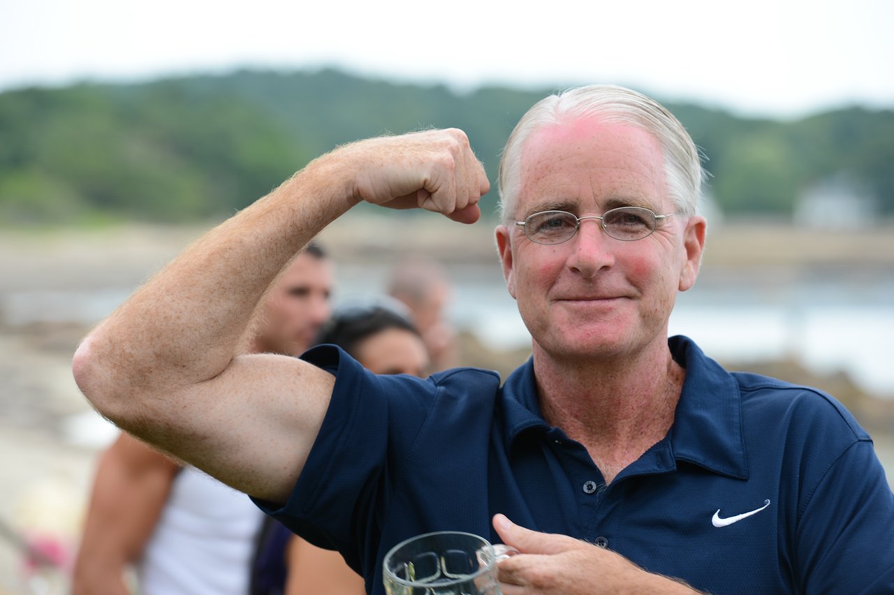 A man in a navy blue shirt flexes his arm and smiles while holding a drink at an outdoor gathering.