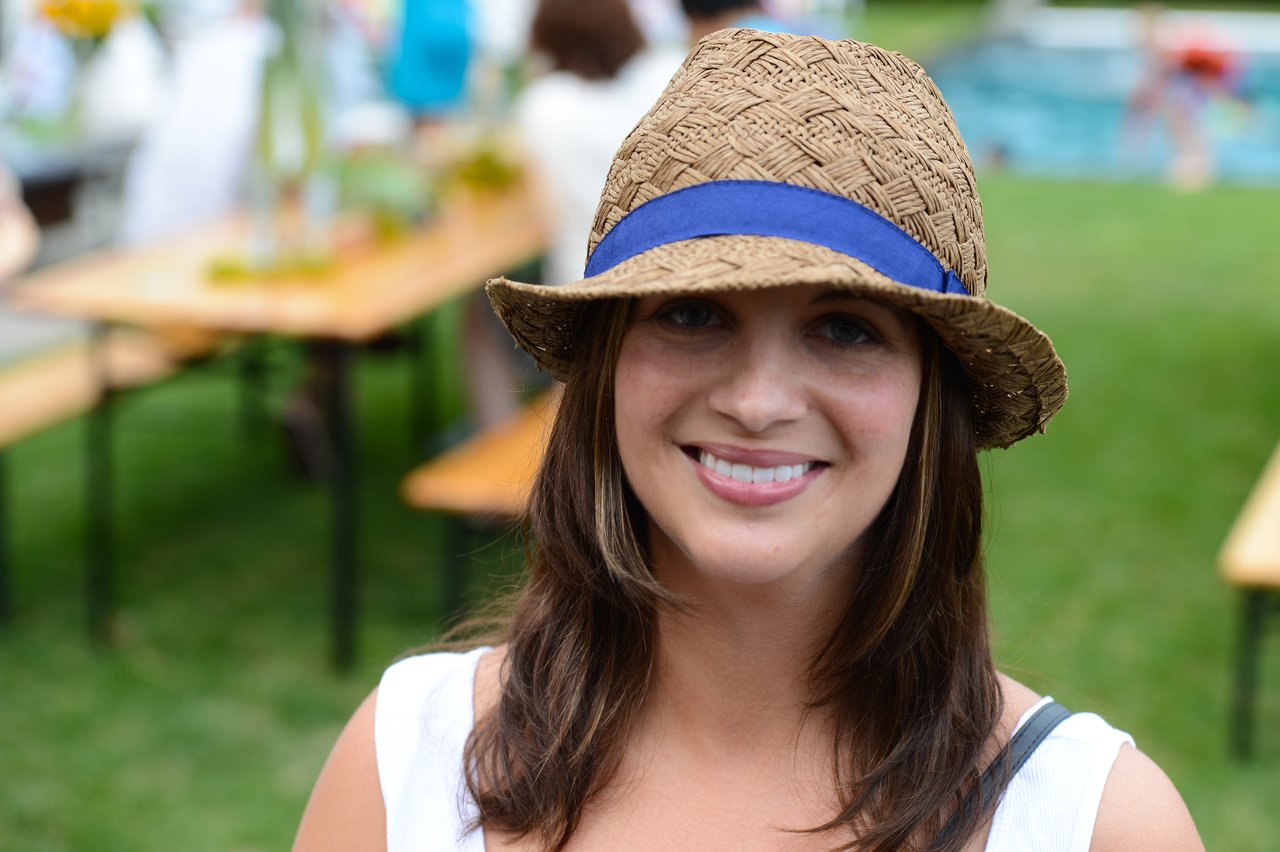 A woman wearing a straw hat with a blue ribbon smiles at the camera at an outdoor party.