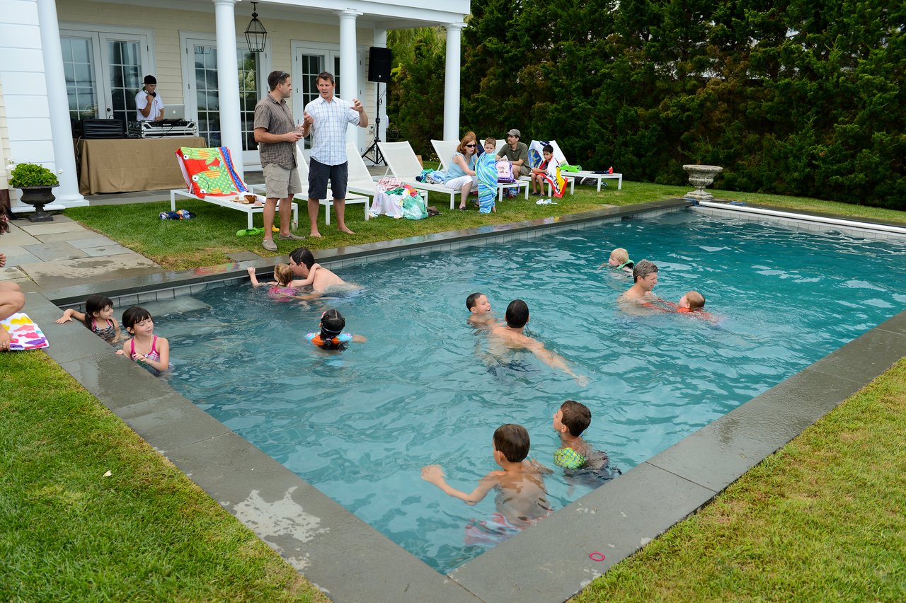 People are swimming in a pool while others relax on lounge chairs and chat near a house.