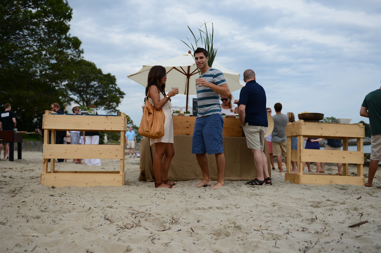 People socializing at a beach party, holding drinks near a wooden bar with food and decorations.