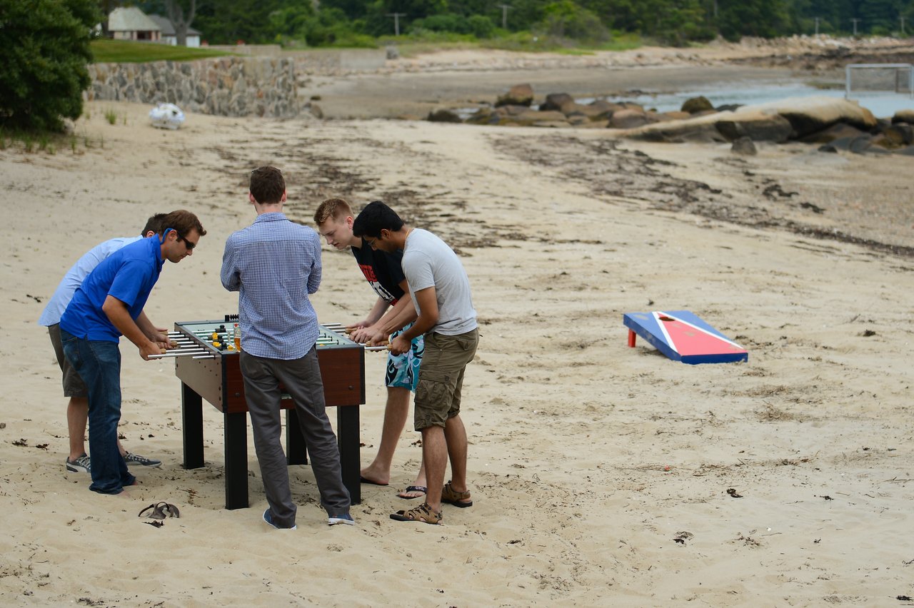 Four men play foosball on a sandy beach while another person watches nearby.