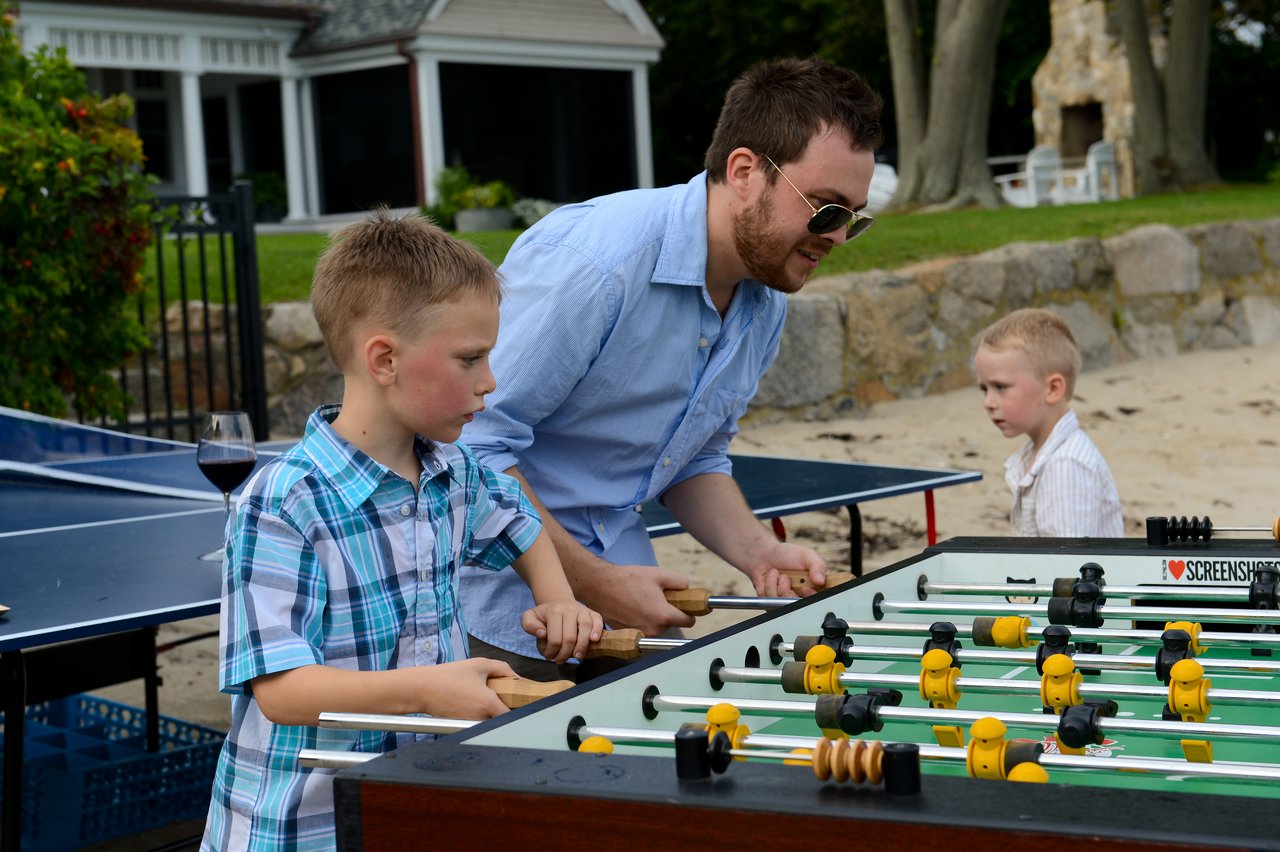 A man and a boy play foosball together outside, while another child watches nearby.