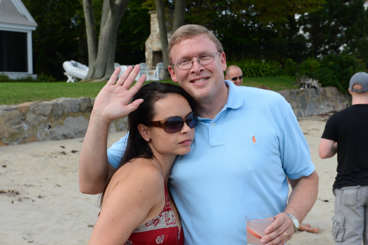 A man in a blue shirt smiles while hugging a woman in sunglasses, who waves at the camera.