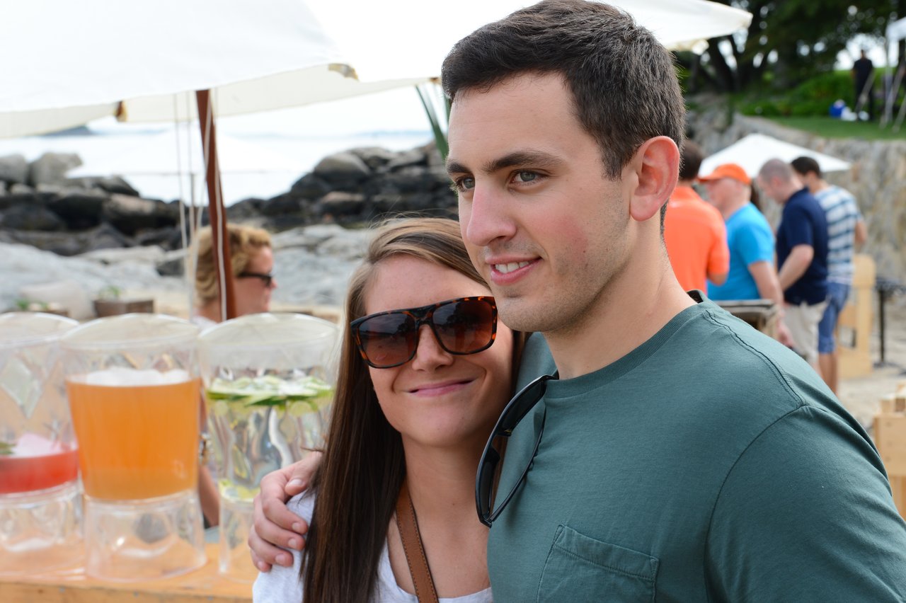 A smiling couple poses together at an outdoor summer party with drinks and other guests in the background.