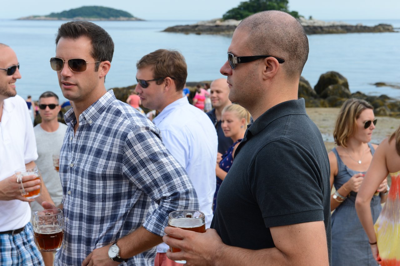 People wearing sunglasses and casual clothes stand on a beach, holding drinks and talking at an outdoor party.