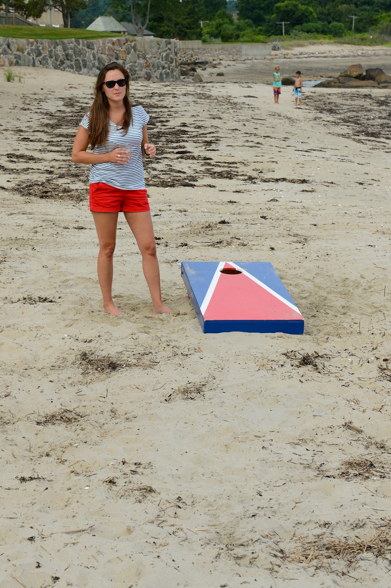 A woman in sunglasses stands on a sandy beach near a cornhole board, holding a drink.