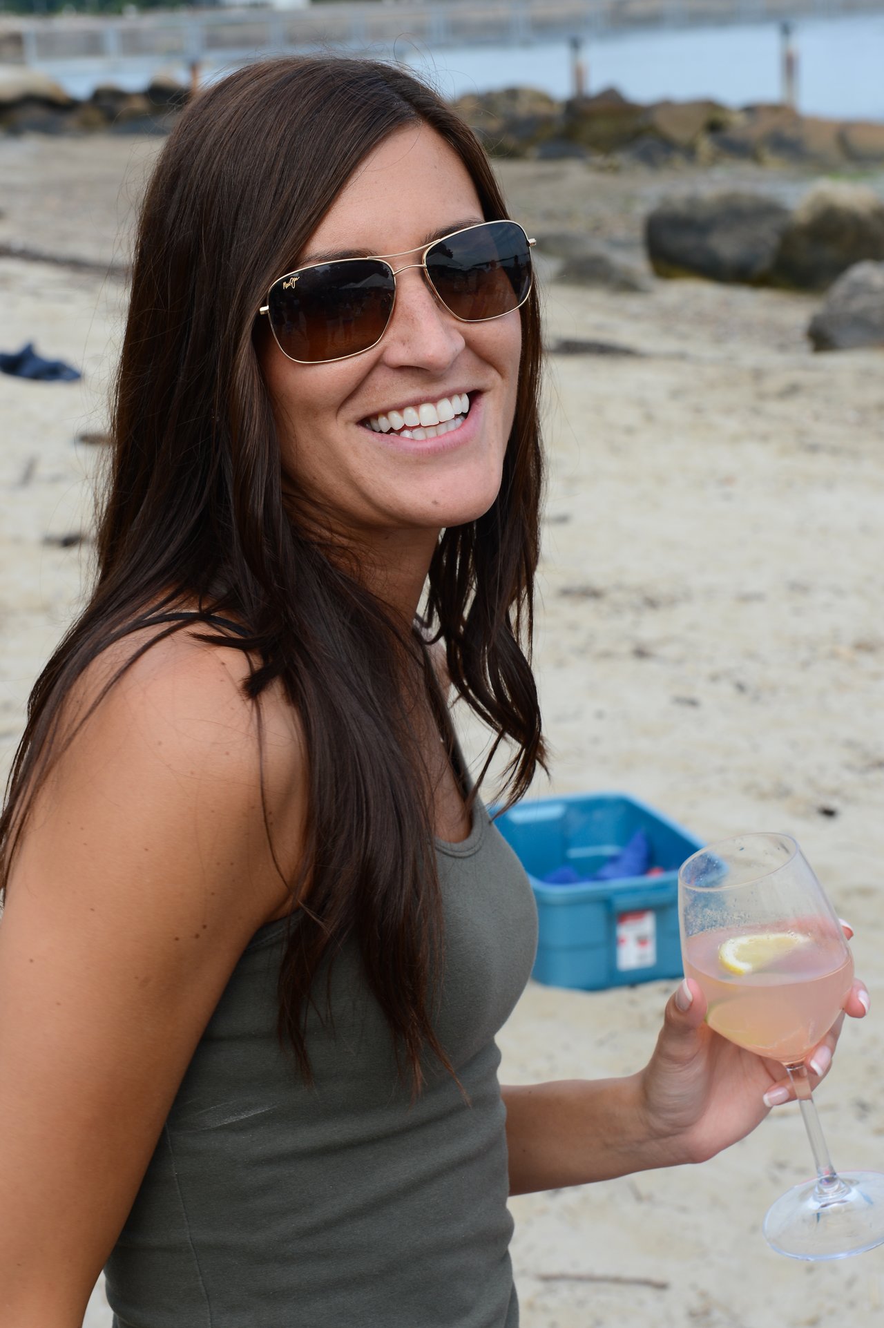 A woman wearing sunglasses smiles while holding a glass of wine at a beach gathering.